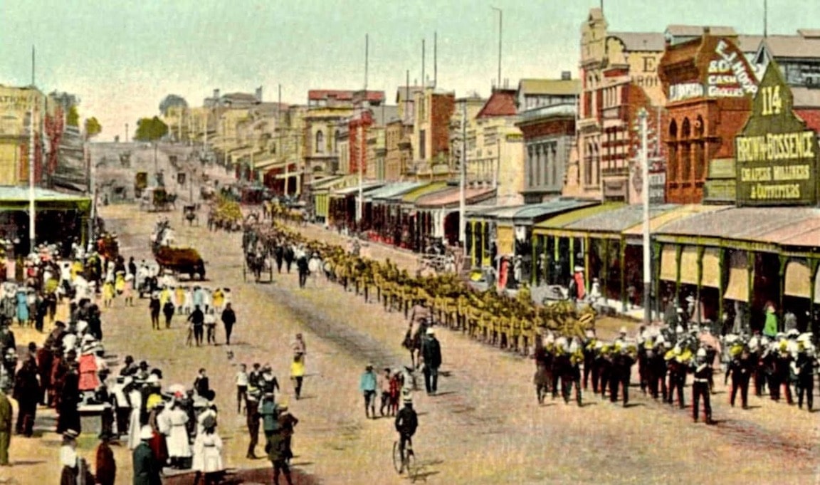 Colourised historical photograph of a military band marching down a busy Geelong street lined with period shopfronts and crowds